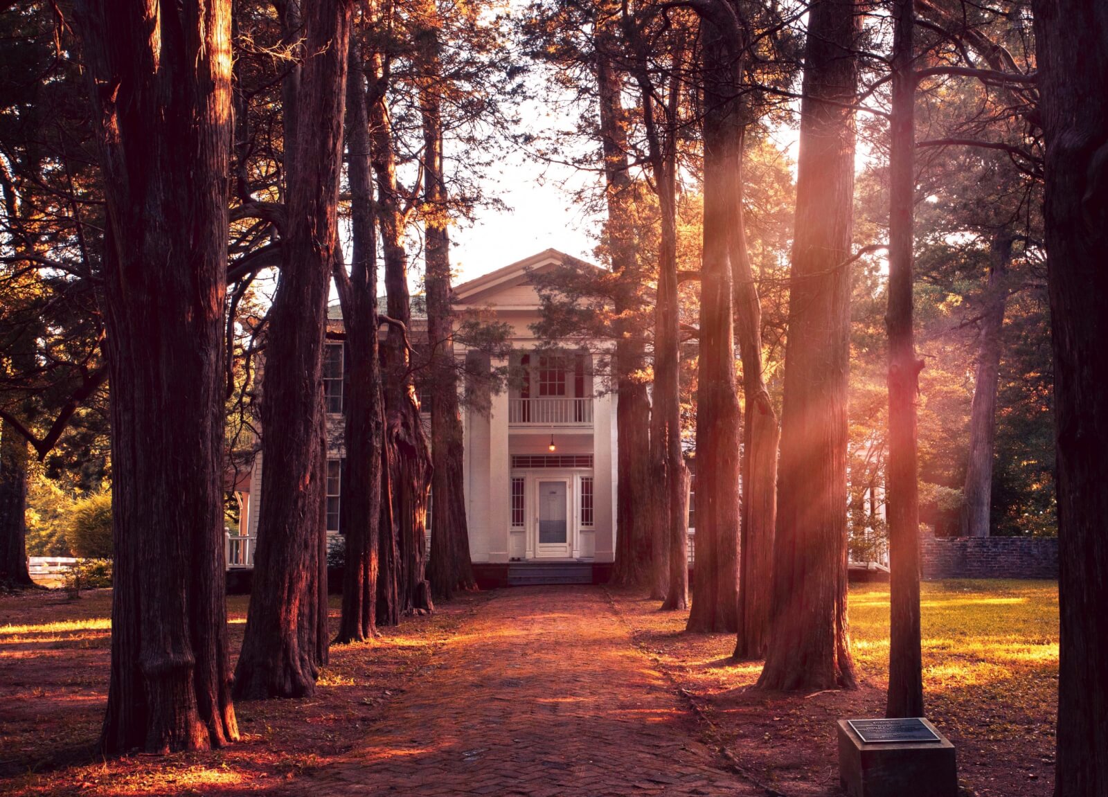A white, columned house sits at the end of a tree-lined brick path in a wooded area, reminiscent of classic Southern charm found in SEC Towns, with sunlight filtering through the tall trees.