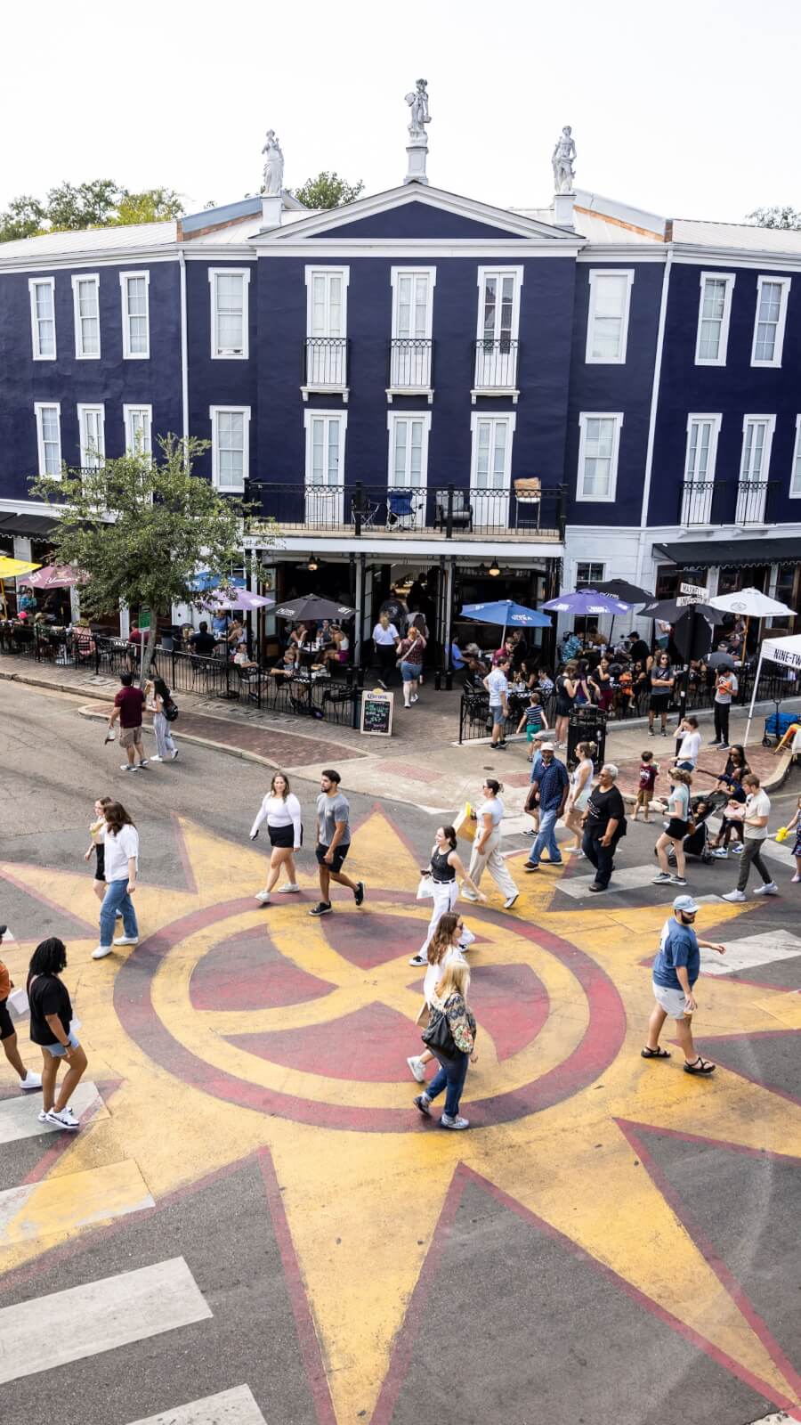 People cross a city street painted with a large sunburst pattern in front of a dark blue building with outdoor restaurant seating, capturing the vibrant atmosphere often found in SEC Towns.