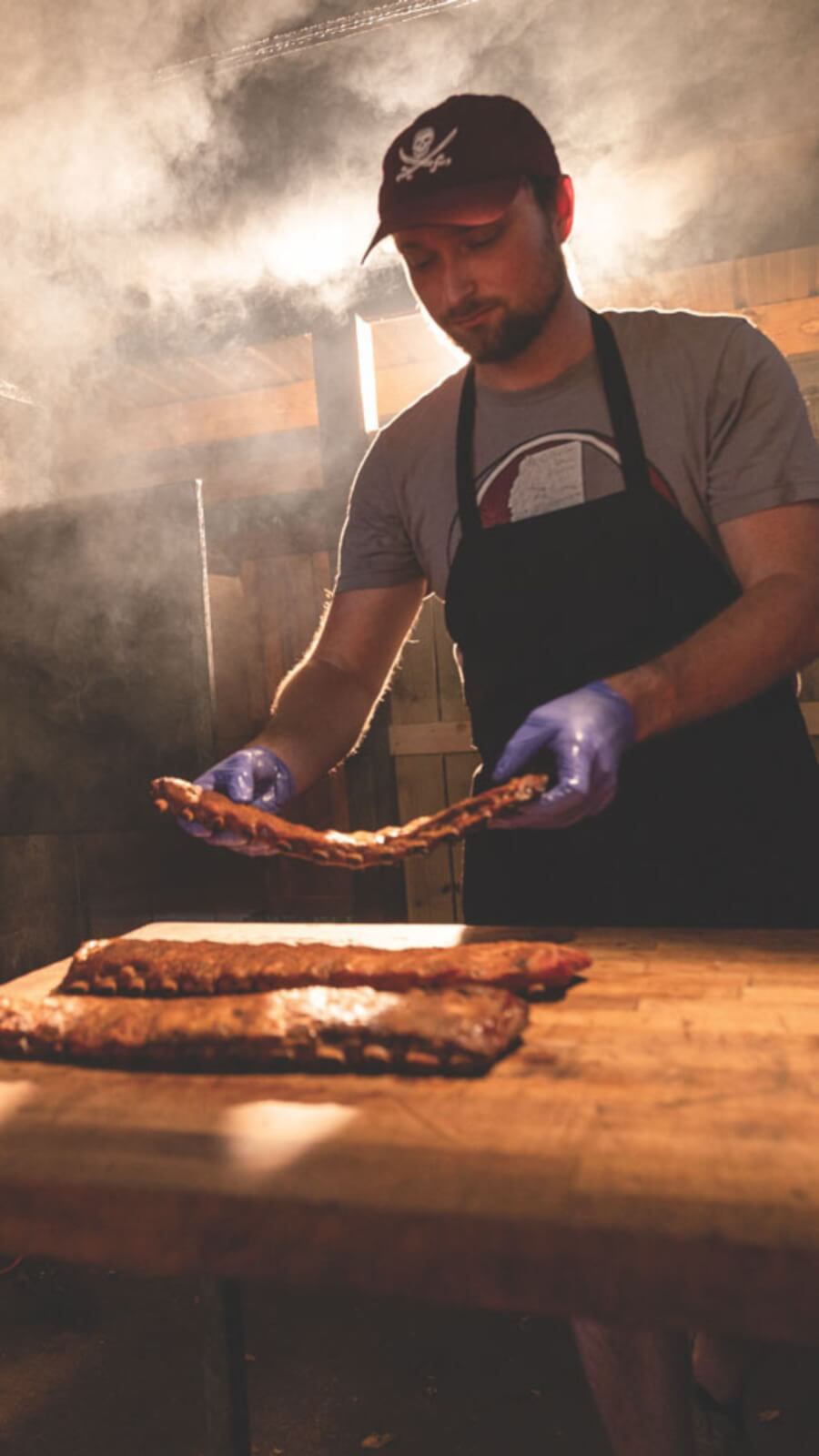 In a smoky setting reminiscent of SEC Towns, a person wearing gloves and an apron places barbecued ribs on a wooden table.
