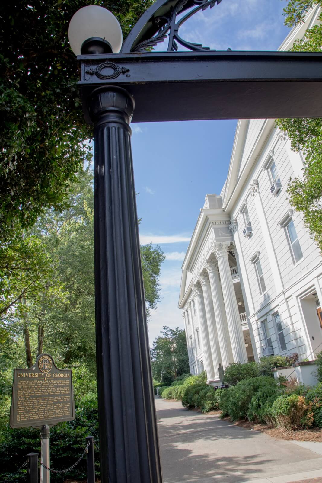 Black iron arch with a lamp in the foreground, a historic marker, and a white columned building at the University of Georgia in the background highlight the charm found in classic SEC Towns.