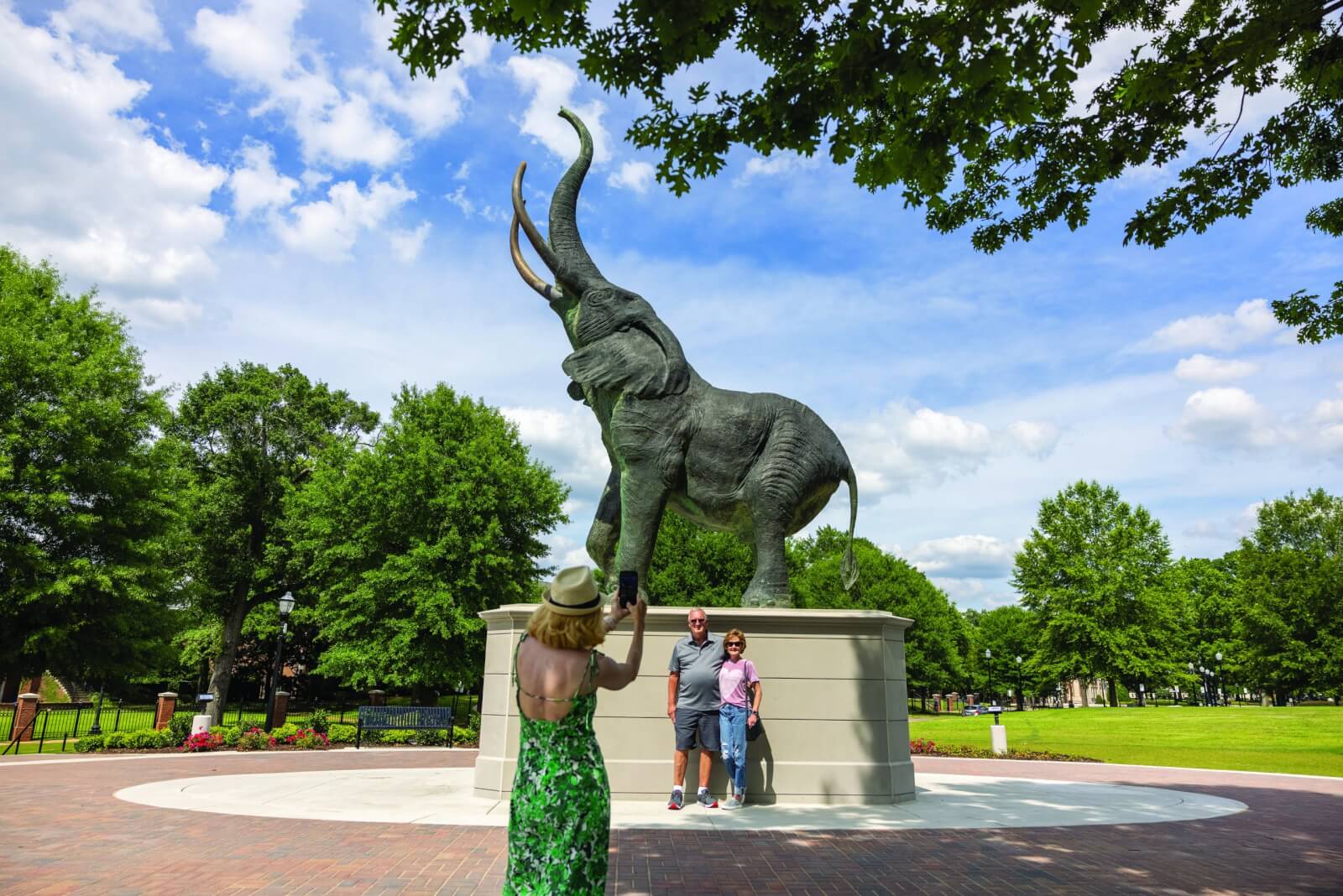 A woman takes a photo of two people standing by a large elephant statue outdoors on a sunny day, capturing the spirited atmosphere typical of SEC Towns.