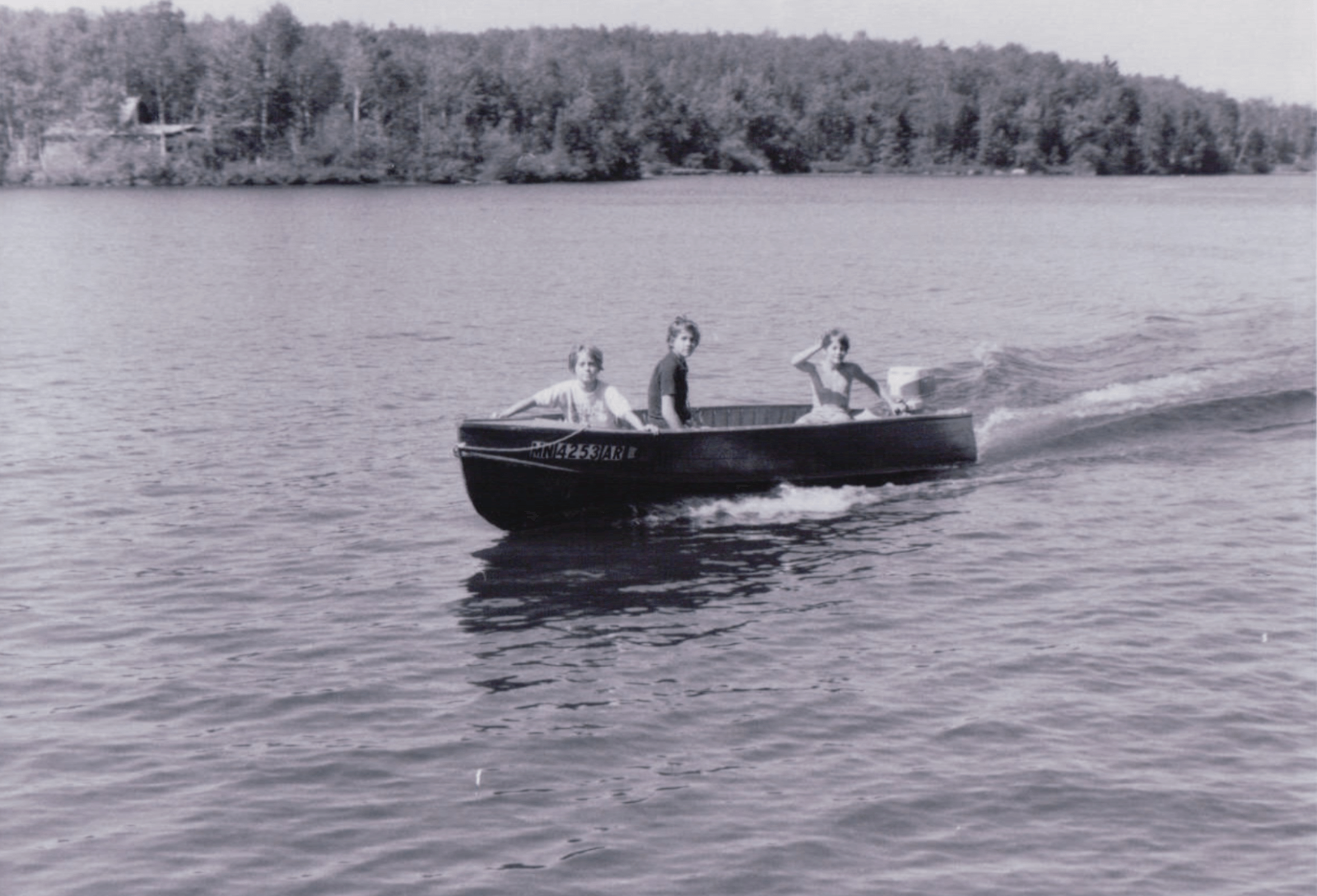 Three people embrace lake life as they ride in a small motorboat on a calm lake, surrounded by trees. The black and white photo captures the peaceful atmosphere.