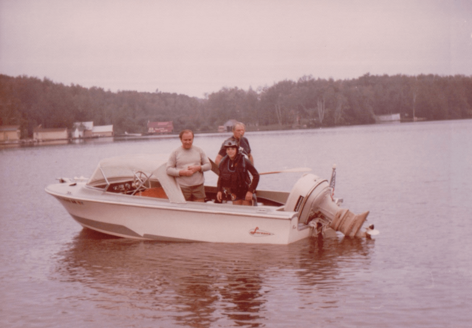 Three people stand on a small white motorboat in calm water, embracing lake life near a wooded shoreline with houses in the background.