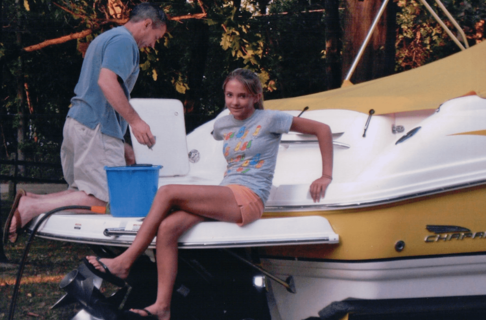 A man stands on a yellow and white boat next to a blue bucket while a girl, enjoying lake life, sits on the back and smiles at the camera. Trees are visible in the background.