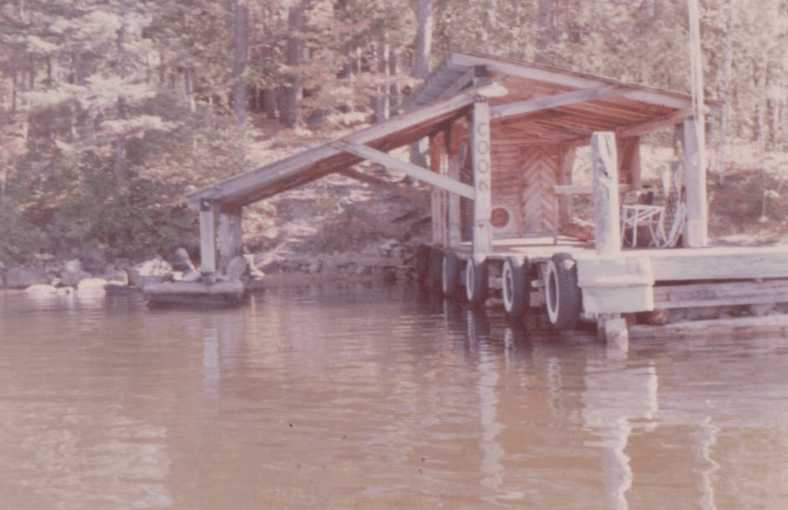 A small wooden boathouse with a slanted roof sits on a dock over calm water, surrounded by trees; tires line the dock as bumpers—a peaceful scene capturing the essence of lake life.