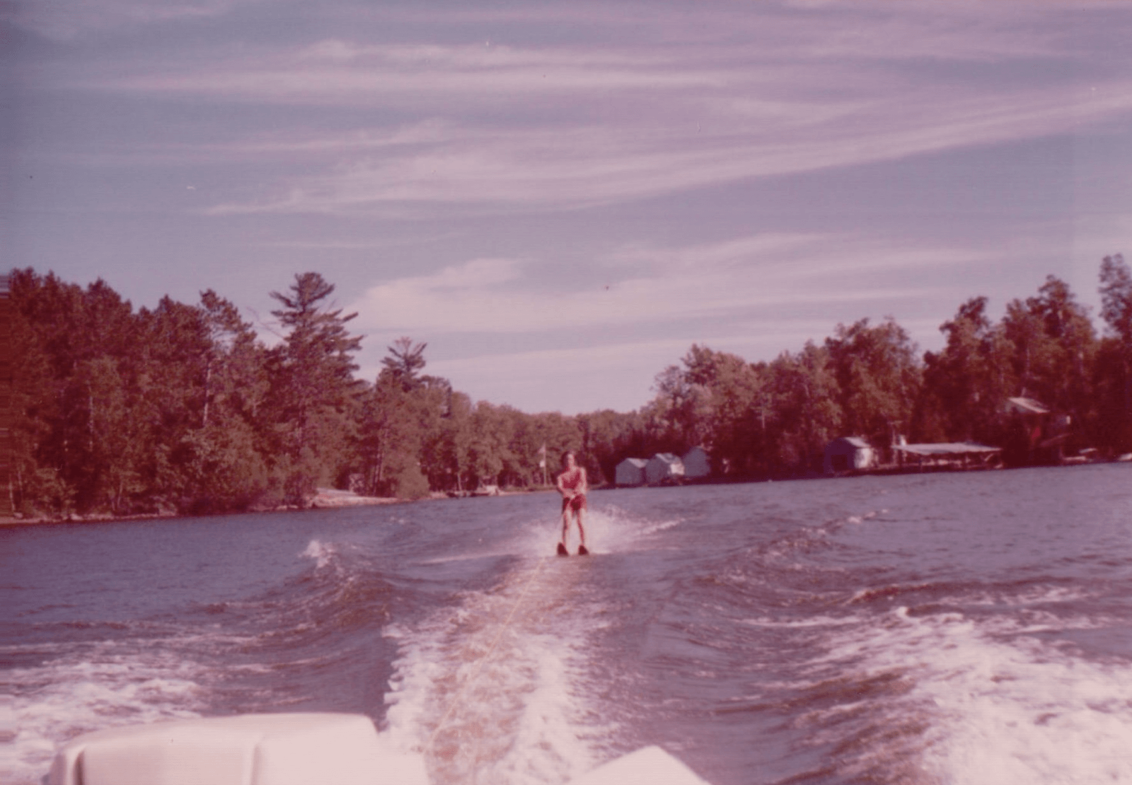 A person is water skiing on a lake, embracing lake life while being pulled by a boat, with trees and several small buildings dotting the shoreline in the background.