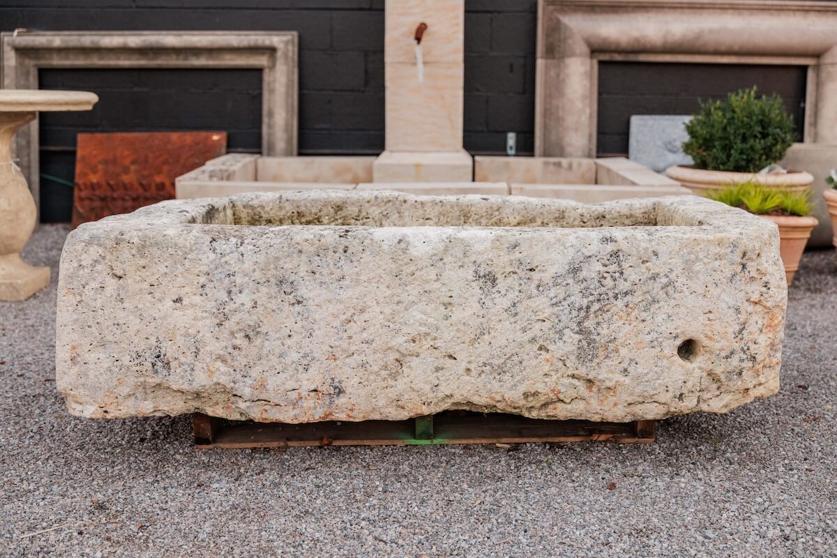 A large rectangular stone trough sits on gravel, with a hole on one side; stone planters and architectural elements complete the scene—a perfect touch for Birmingham luxe loves garden decor.