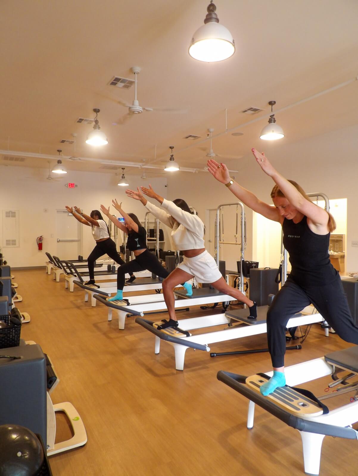 Four people exercise on reformer machines in a brightly lit Pilates studio in Cahaba Heights, each balancing with arms raised while facing forward.