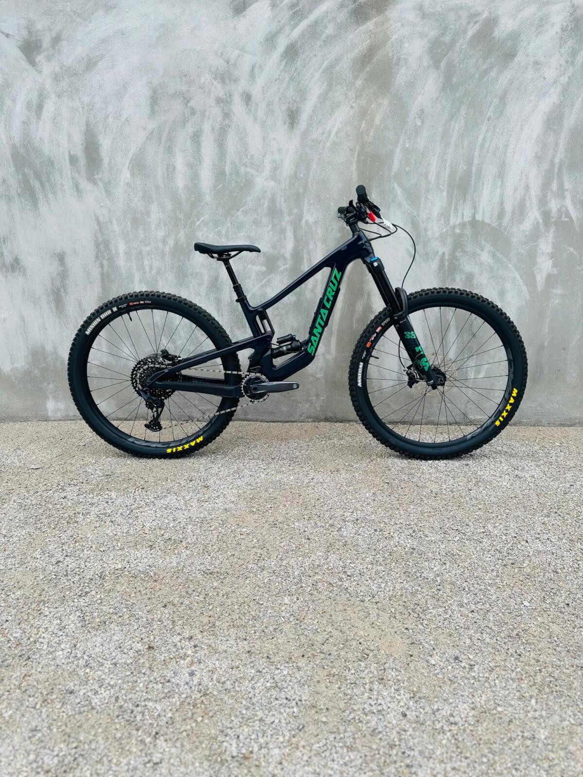 A full-suspension mountain bike with a black frame and green accents is parked against a textured concrete wall on a gravel surface, capturing the adventurous spirit of Cahaba Heights where you can eat, shop, and explore outdoor trails.