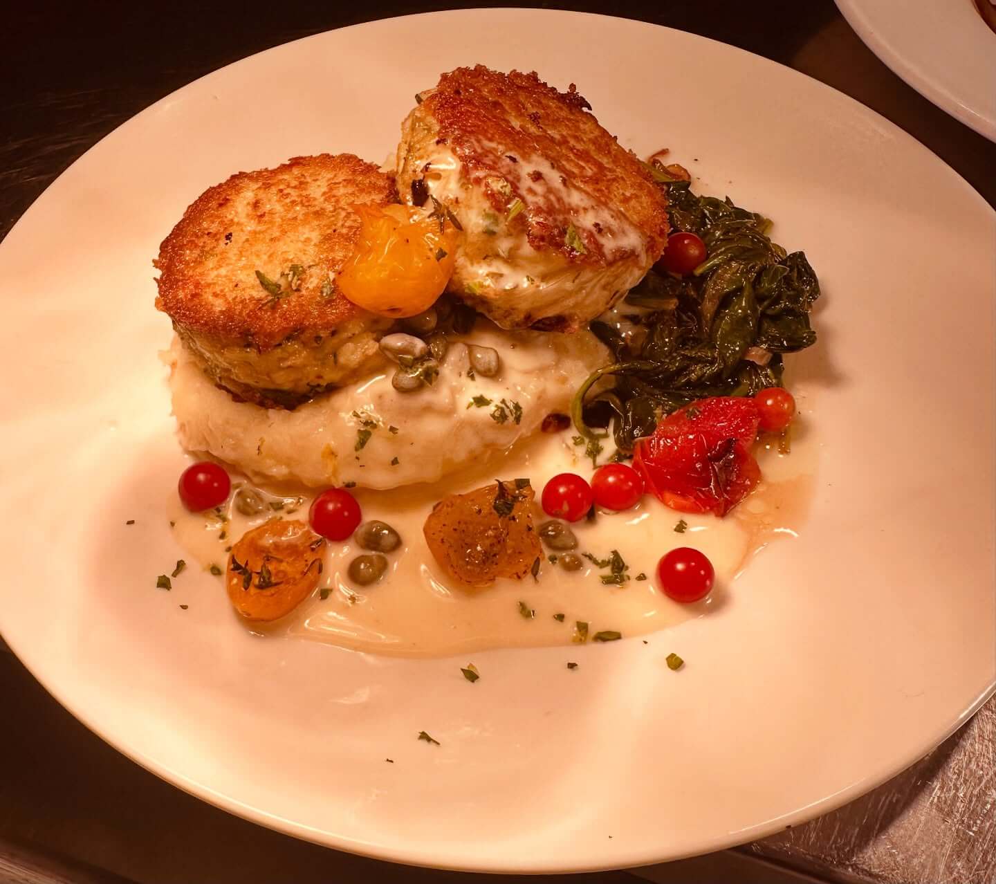 Two crab cakes served over mashed potatoes with savory sauce, cherry tomatoes, capers, and sautéed greens.