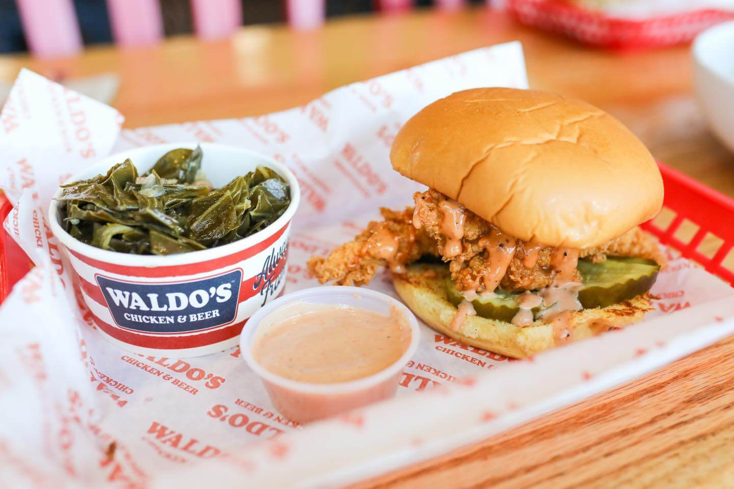 Enjoy a fried chicken sandwich with pickles and sauce, served on a tray with cooked greens and dipping sauce—perfect to eat after you shop in Cahaba Heights.