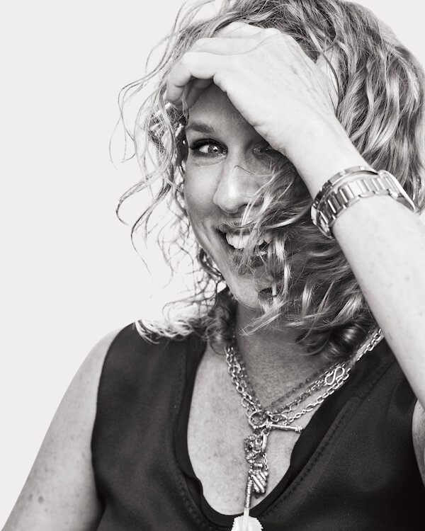 Meet Birmingham Travel & Lifestyle Writer Christiana Roussel - 4 A woman with curly hair smiles, partially covering her face with one hand. She wears layered necklaces, a sleeveless top, and a watch. This black and white image captures the effortless style of Christiana Roussel.