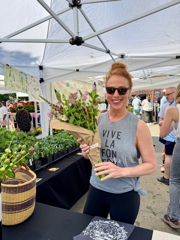 Meet Birmingham Travel & Lifestyle Writer Christiana Roussel - 5 A woman wearing sunglasses and a "VIVE LA FON FON" shirt holds a bouquet of flowers at an outdoor market stall under a canopy.