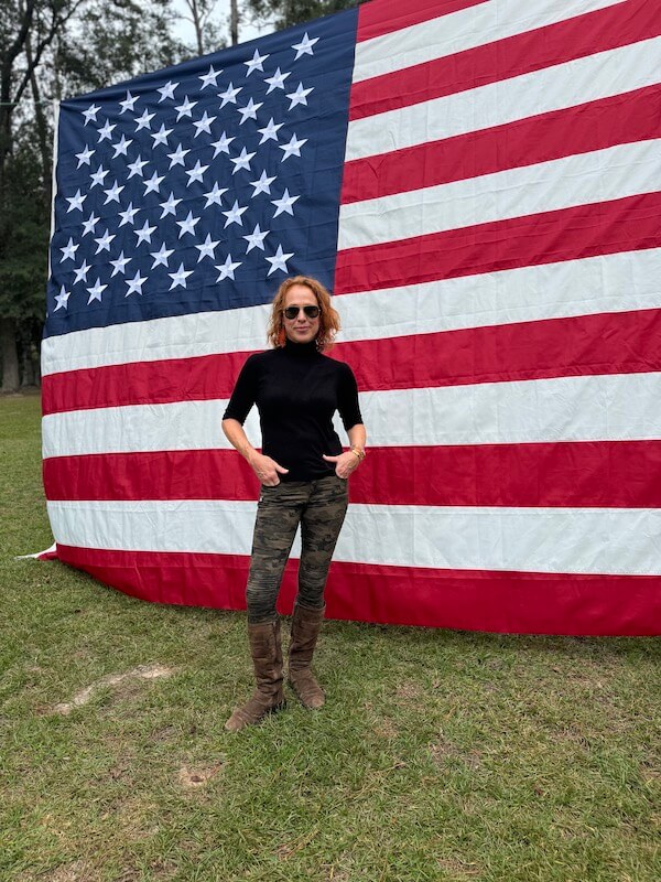 Meet Birmingham Travel & Lifestyle Writer Christiana Roussel - 6 A woman wearing sunglasses, a black top, camouflage pants, and brown boots stands on grass in front of a large American flag.