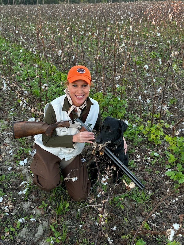 Meet Birmingham Travel & Lifestyle Writer Christiana Roussel - 2 A woman in hunting attire kneels in a field beside a black dog, holding a shotgun and smiling for the camera.