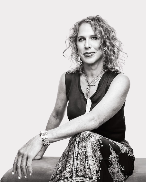 Meet Birmingham Travel & Lifestyle Writer Christiana Roussel - 1 Christiana Roussel, with her curly hair, sits on a bench in a black and white photo, wearing a sleeveless top, patterned skirt, necklace, and bracelets, her arm resting casually on her knee.