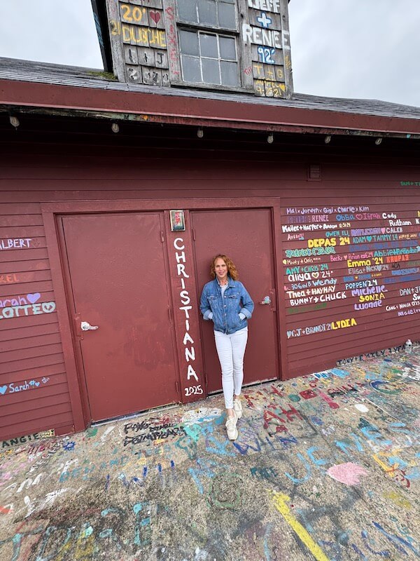 Meet Birmingham Travel & Lifestyle Writer Christiana Roussel - 3 A person in a blue denim jacket and white pants stands in front of a red building covered with colorful names and graffiti.