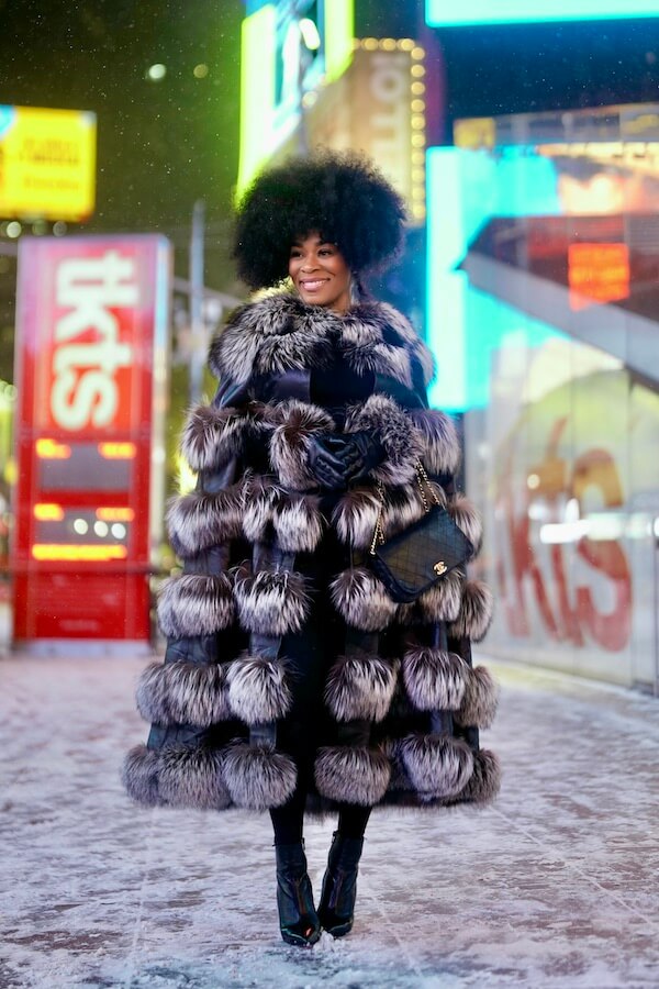 A woman in a large, multi-layered fur coat stands outdoors on a snowy street at night, surrounded by bright neon signs.