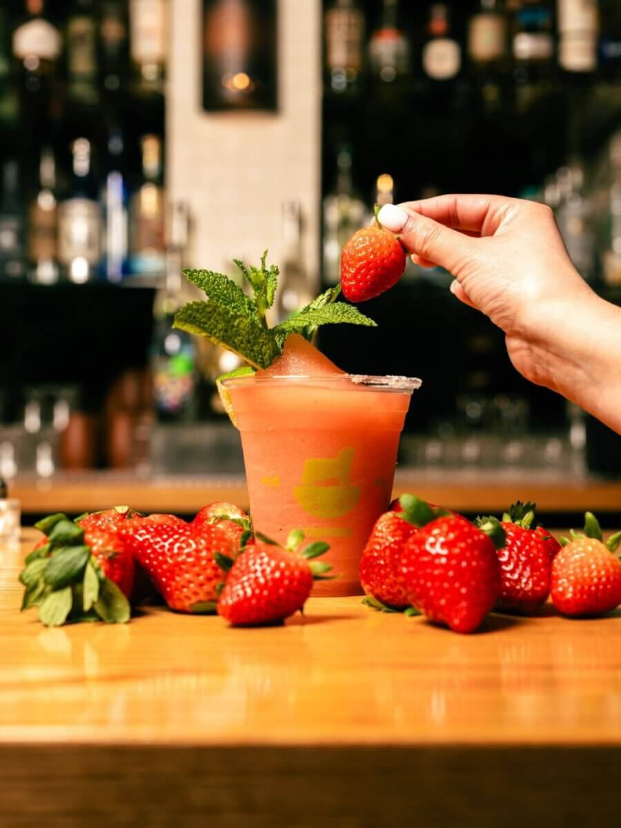 10 Frozen Cocktails to Try in BHAM This Summer - 11 A hand holds a strawberry above a frozen cocktail garnished with mint, surrounded by fresh strawberries on a bar counter. Bottles are visible in the background.