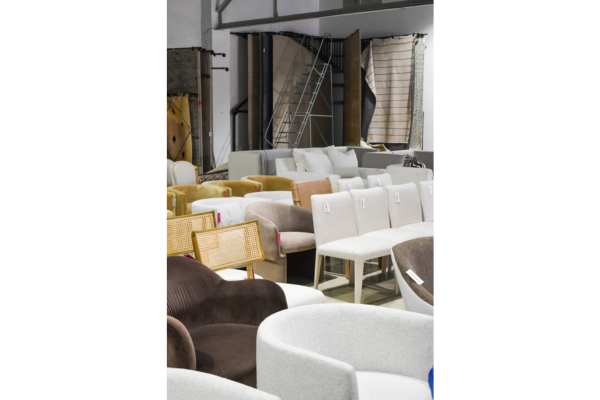 Rows of assorted upholstered chairs and sofas are displayed in a showroom, with stacked rugs and a ladder visible in the background, ready for discovery at our Once-A-Month Shopping Event.