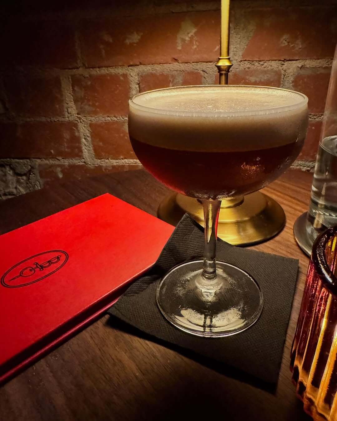 A cocktail in a coupe glass sits on a black napkin beside a red menu, a lamp, and a glass of water on a wooden table against a brick wall at Bygones Bar in The Battery.