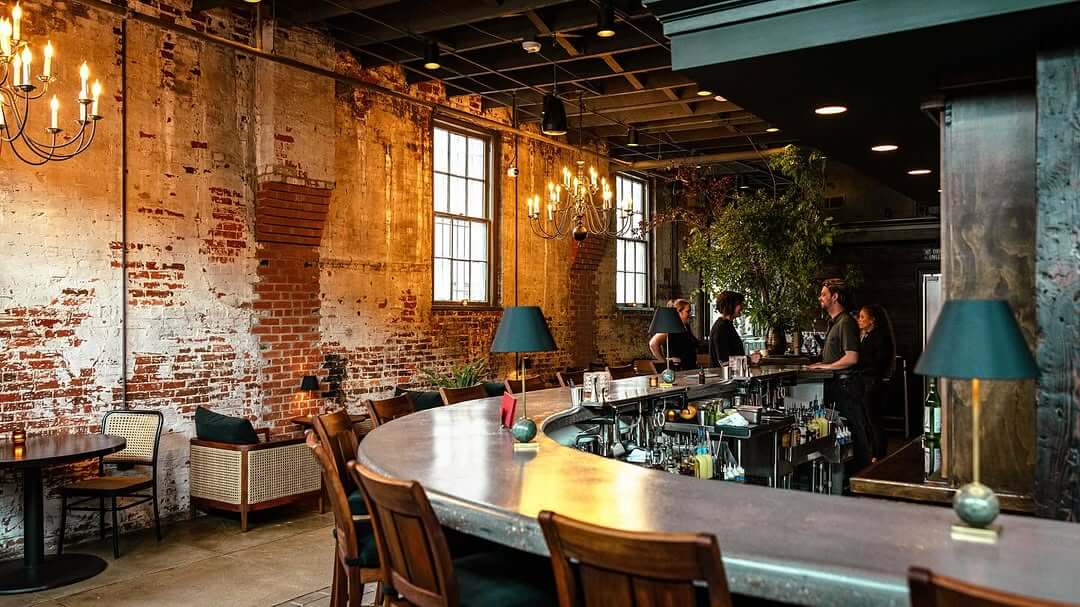 A curved bar with wooden stools in an industrial-style space at Bygones Bar, featuring exposed brick walls, chandeliers, large windows, and several people behind the counter.