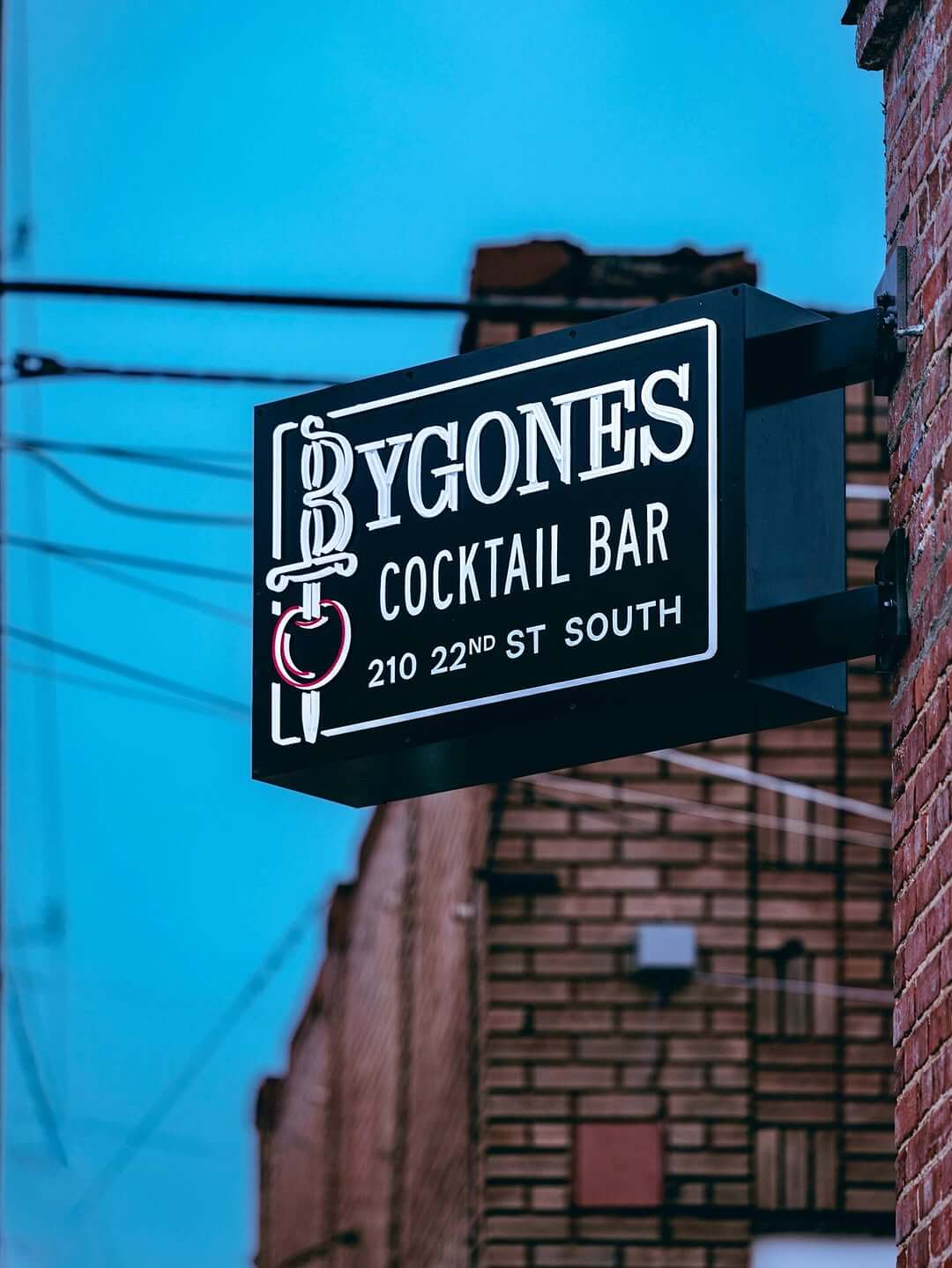A black sign for Bygones Cocktail Bar with white lettering and red accents hangs on a brick wall at 210 22nd St South, just steps from The Battery II.