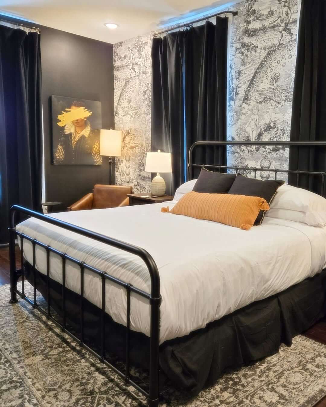 A bedroom features a black metal bed with white bedding and an orange accent pillow, patterned wallpaper at The Painted Lady Hotel, dark curtains, a rug, leather chair, lamps, and wall art.