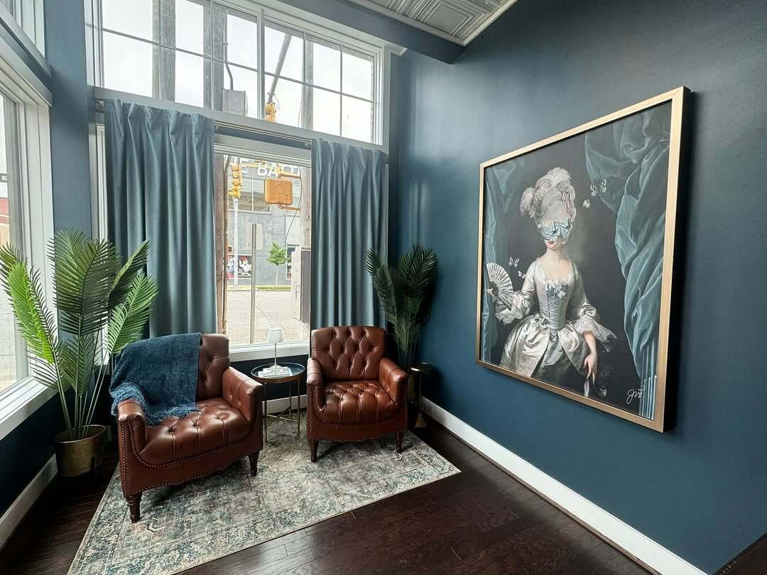 Two brown leather armchairs sit by a window with blue curtains at the Painted Lady Hotel, a small table and potted plants nearby, beneath a large framed portrait of a woman in historical attire on a blue wall.