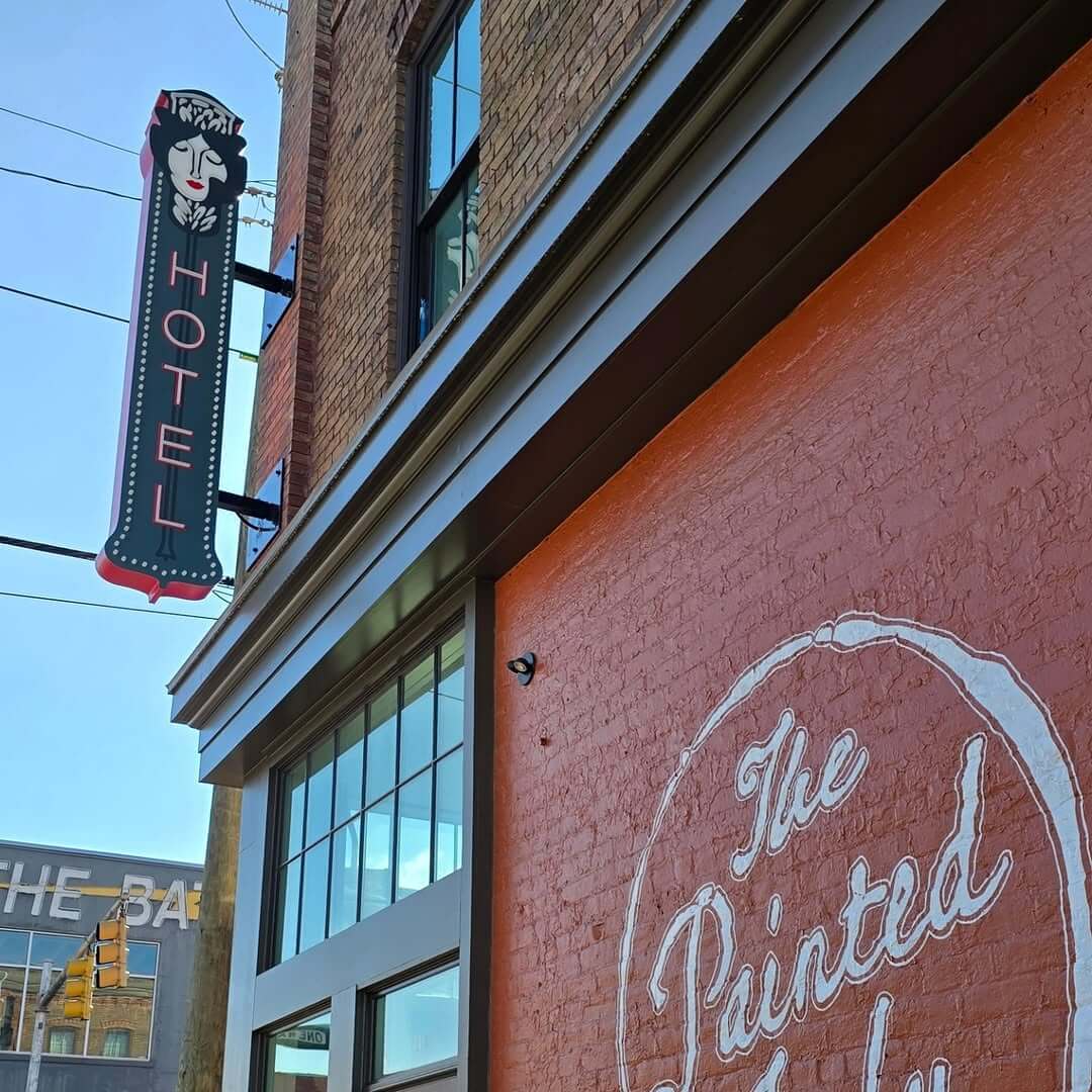 A building with a painted orange brick wall reading "The Painted Lady Hotel" and a vertical neon sign above showing "HOTEL" next to an illustrated woman’s face, located steps from Bygones Bar and The Battery.