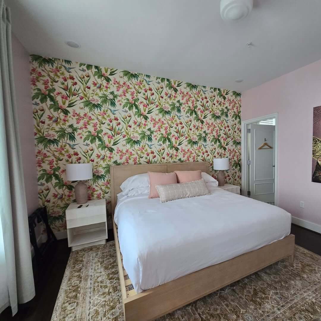 A bedroom with a large bed, white bedding, two nightstands with lamps, floral wallpaper behind the bed, and light pink walls.