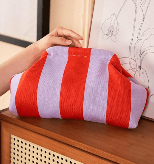 A hand holds a soft, red and lavender Cabana Stripes clutch bag resting on a wooden surface near a botanical line drawing.