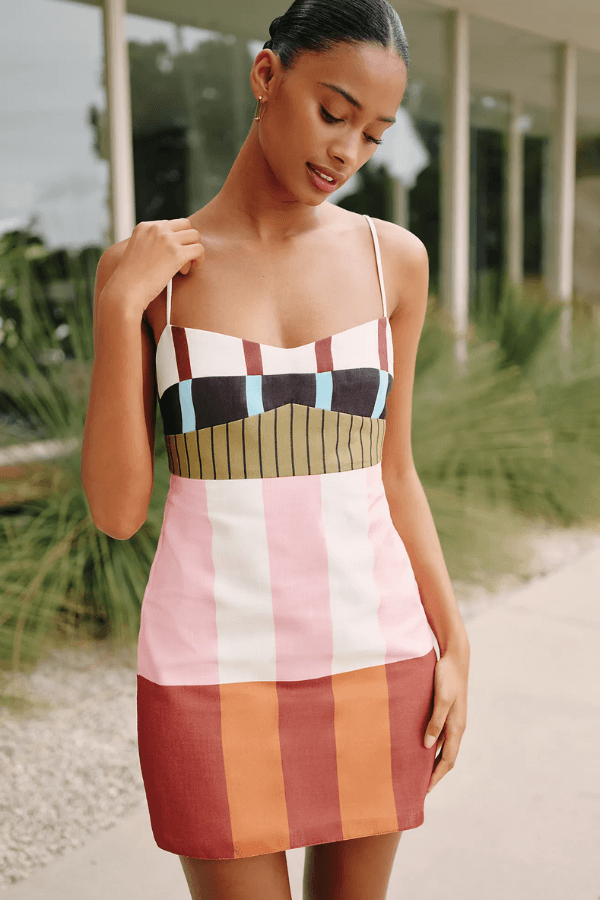 Woman wearing a sleeveless, multicolored mini dress with Cabana Stripes and geometric patterns stands outdoors near a building with large windows and greenery.