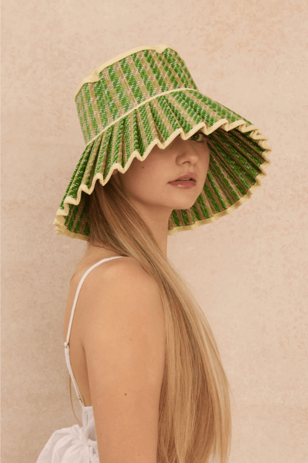 A woman with long blonde hair in a white dress wears a wide-brimmed hat woven with green and yellow cabana stripes, partially covering her eyes, against a beige textured background.