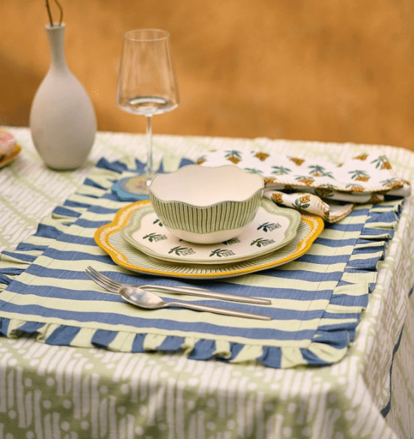 A table set with cabana stripes placemats, patterned plates, a bowl, a fork and knife, a wine glass, a white vase, and a folded napkin with a green and yellow design.
