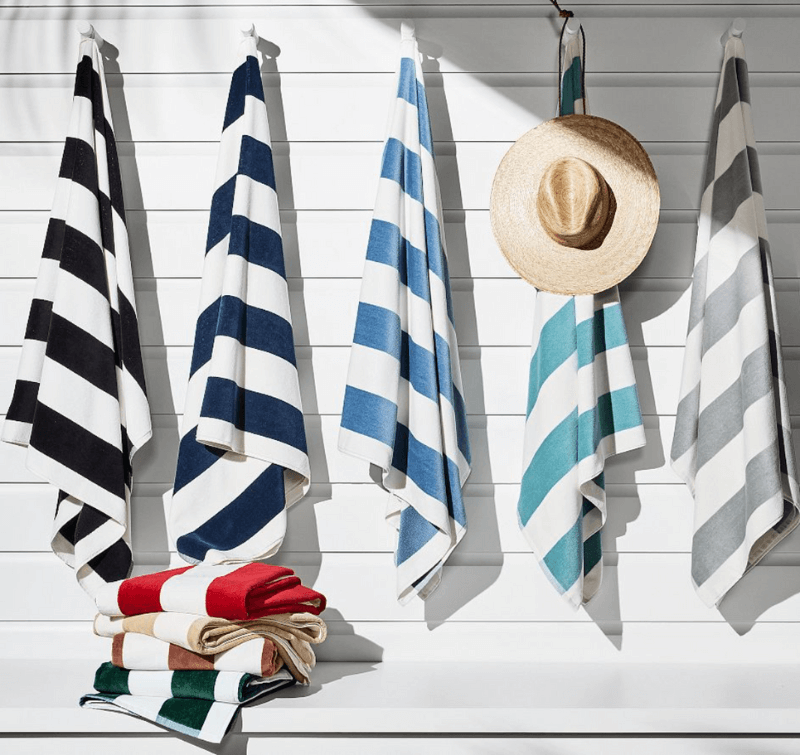 Five Cabana Stripes beach towels hang on hooks, with a straw hat on one hook. A stack of folded towels rests on a white bench below.