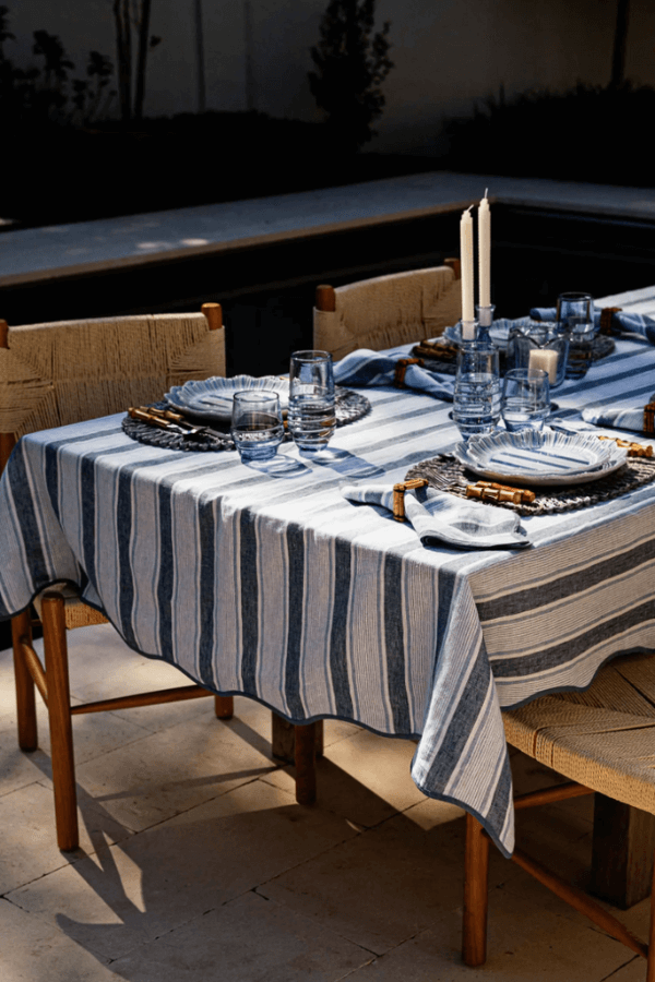 A dining table set outdoors with a blue and white cabana stripes tablecloth, glassware, plates, napkins, woven placemats, and two white candles in holders.