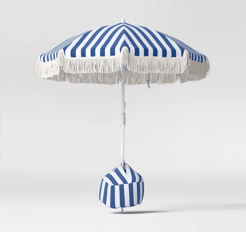 A blue and white Cabana Stripes beach umbrella with white fringe trim stands upright, anchored by a matching striped weighted base, against a plain light background.