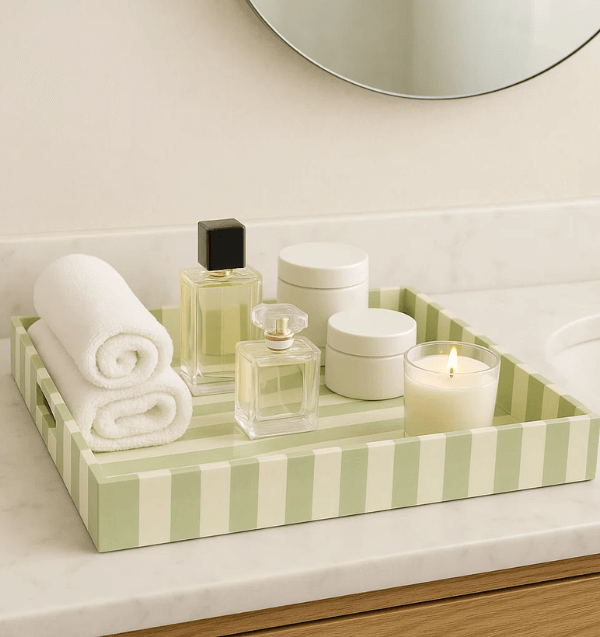 A tray with bold cabana stripes on a bathroom counter holds two rolled towels, two perfume bottles, two jars, and a lit candle.
