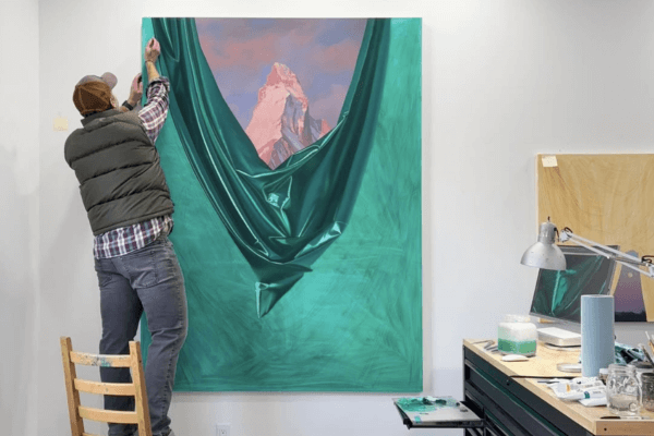 A person stands on a chair, working on a large Shawn Huckins painting of a snow-capped mountain partially revealed behind a green, draped fabric effect in an art studio—an imaginative INTERVENTION with classic landscapes.