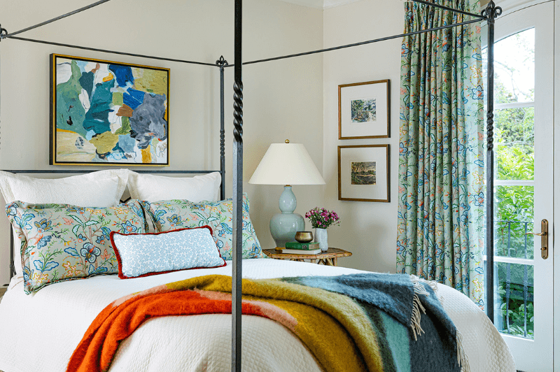 Bright bedroom with a canopy bed, colorful blanket, floral pillows, and a lamp—abstract and landscape art line the walls. A window with patterned curtains overlooks greenery.