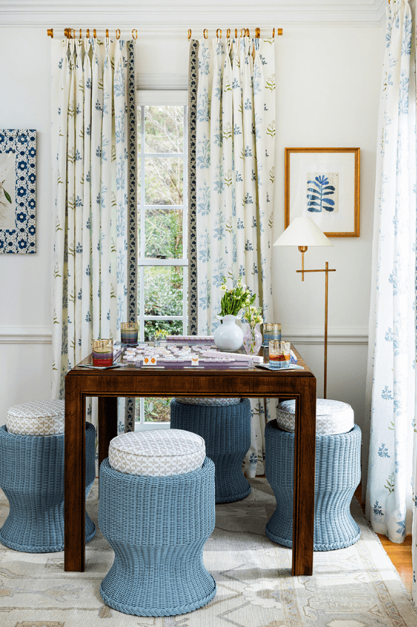 A wooden table with a board game, drinks, and Lena Pizza is surrounded by six blue wicker stools in a bright room with floral curtains, a floor lamp, and framed wall art.