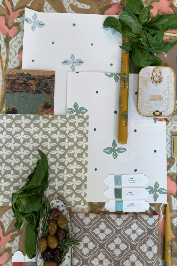 A flat lay of patterned papers, a postcard, canned sardines, fresh basil, green and black olives, embroidery floss, and a patterned fabric on a floral background evokes artisanal charm.