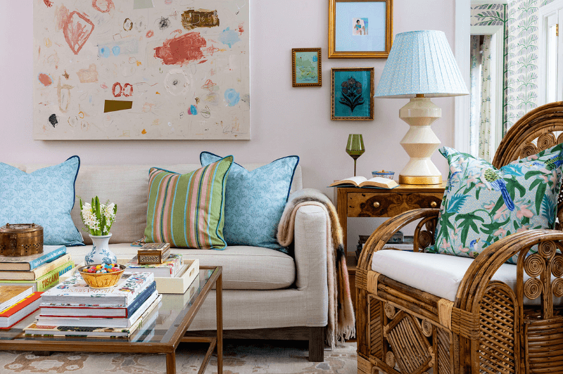 A colorful living room with a beige sofa, blue and striped pillows, a wicker chair, a glass coffee table with books set beneath abstract artwork on a pale wall.