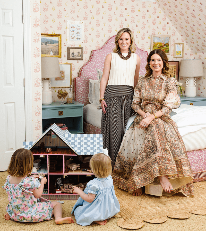 Two women pose in a decorated bedroom while two young girls in blue dresses play with a dollhouse on the floor.