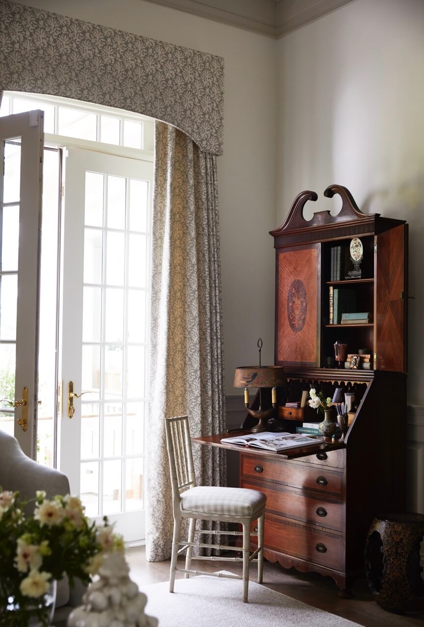 A traditional wooden secretary desk with an open writing surface and shelves stands beside a window with patterned curtains.