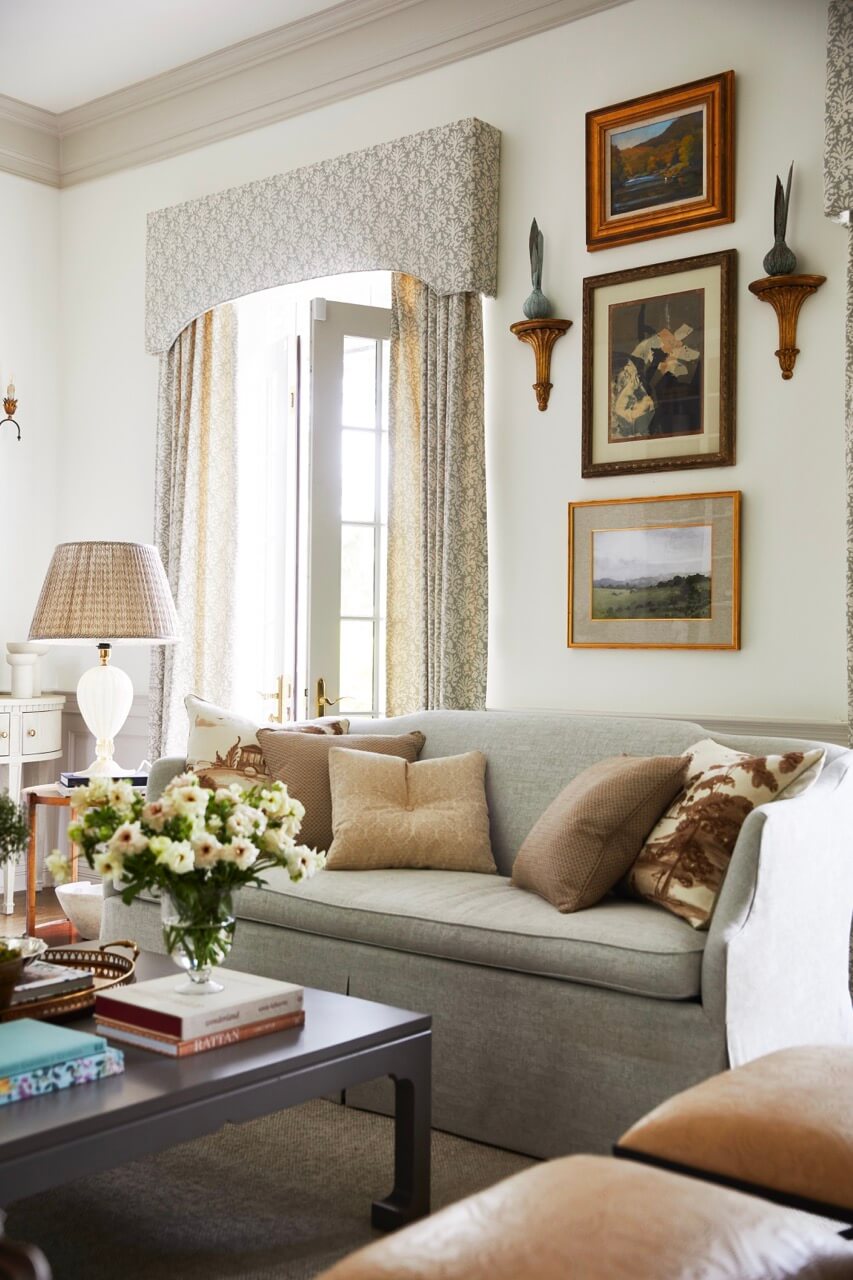 A softly lit living room with a light gray sofa, neutral pillows, framed wall art, a coffee table with books and flowers, and French doors with patterned curtains captures the lakeside beauty of a South Carolina retreat.