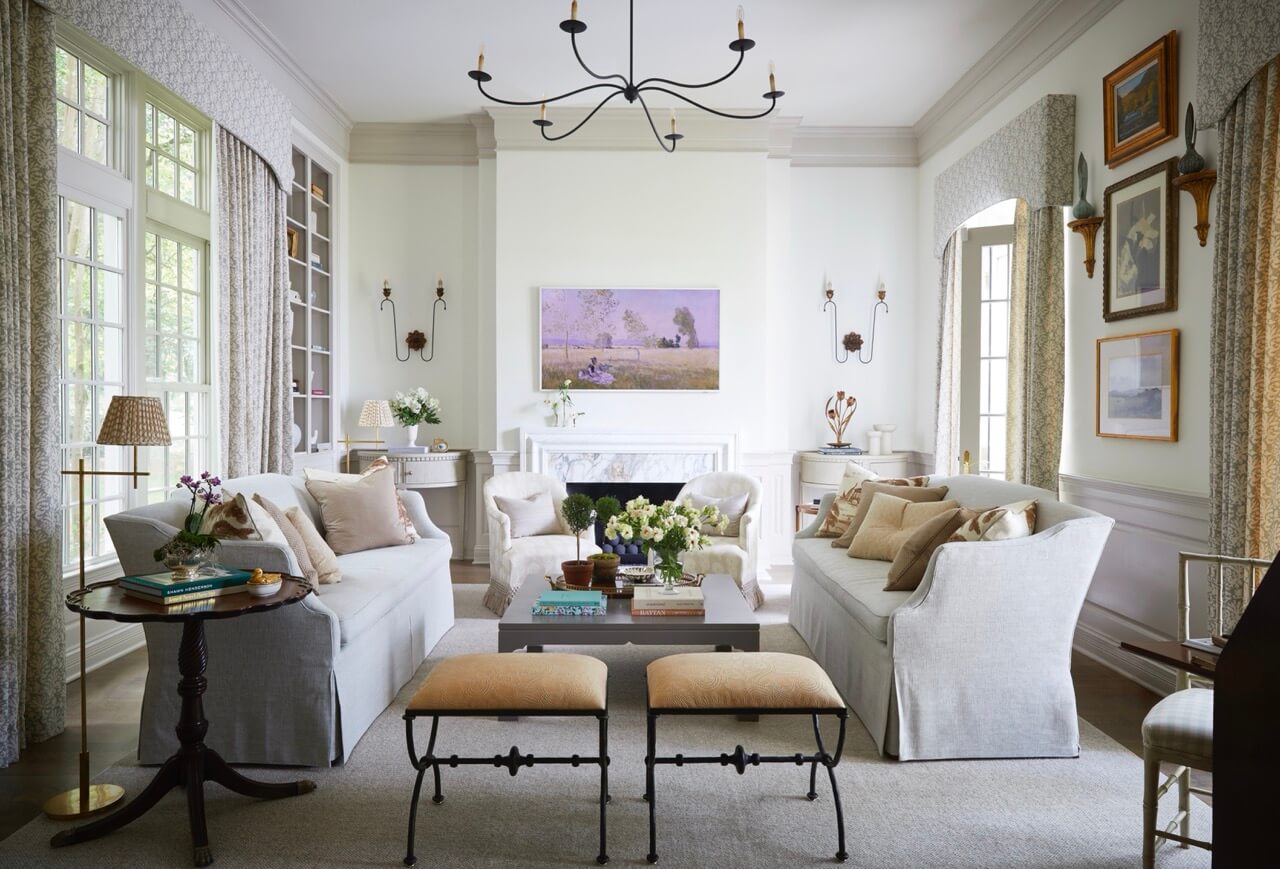 A bright living room with two sofas, armchairs, a fireplace, and large windows captures lakeside beauty. Wall art, books, lamps, and flowers decorate the space in neutral tones at this South Carolina retreat.