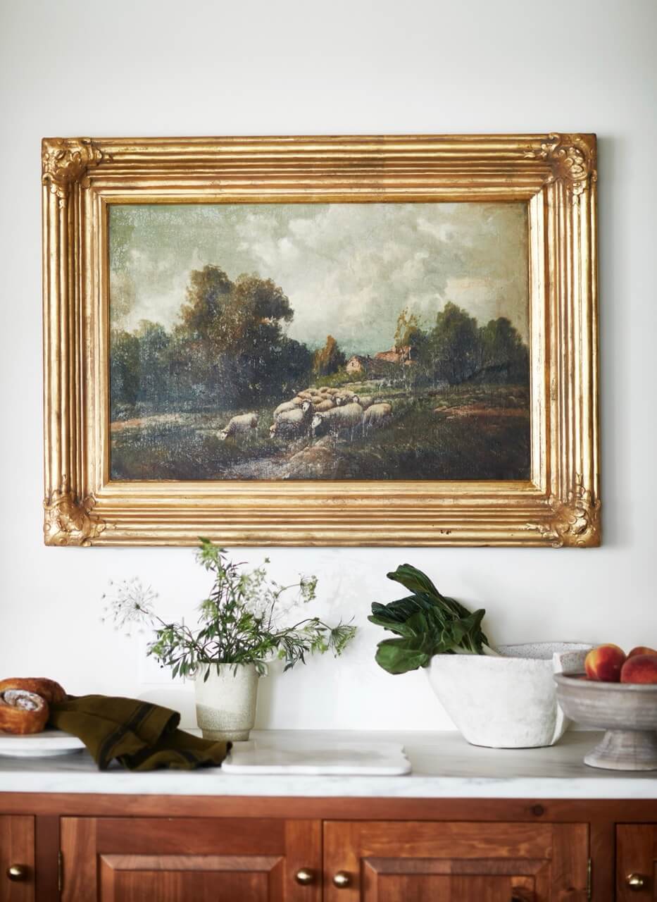 A gold-framed landscape painting hangs above a marble countertop with a potted plant, a loaf of bread, leafy greens in a bowl, and peaches on a wooden sideboard.