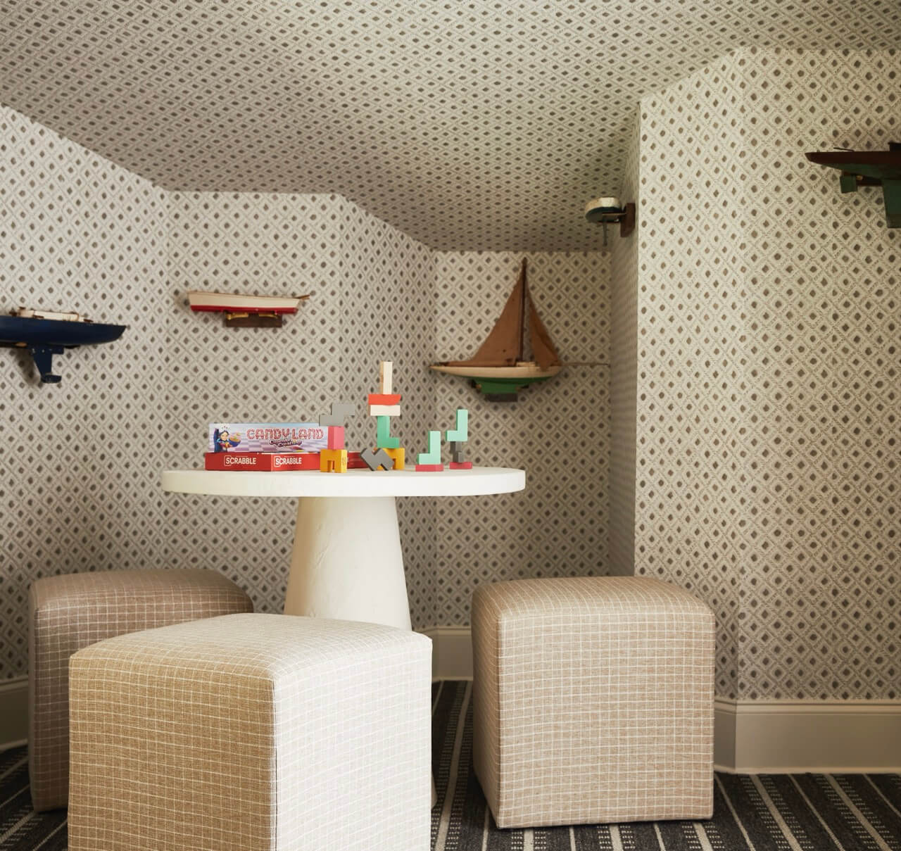 Children’s playroom with patterned wallpaper, a round white table, two plaid ottomans, board games, wooden blocks, and small shelves displaying toys and a model sailboat.